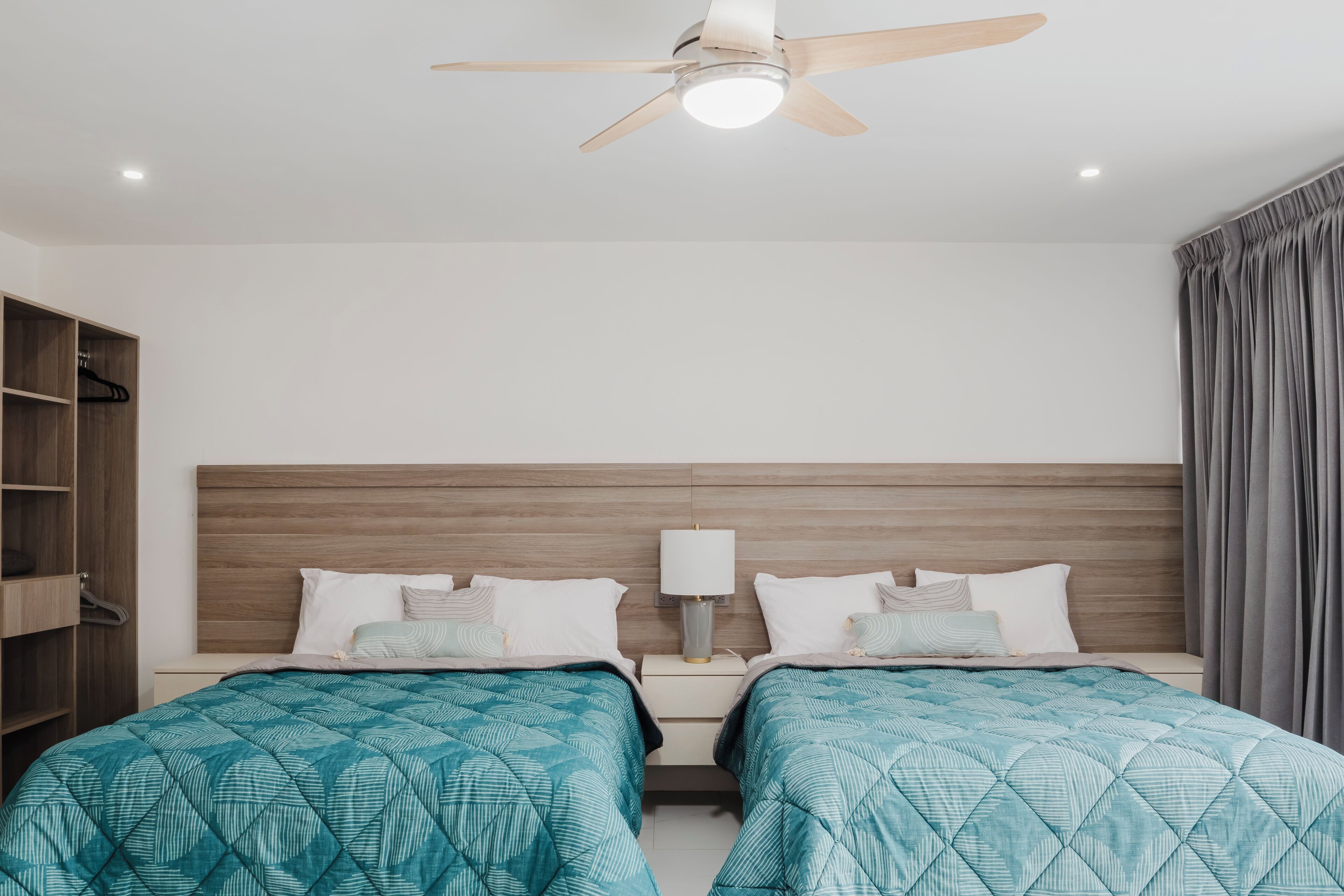 Une chambre d’hôtel avec deux lits, une tête de lit en bois, un ventilateur de plafond, une table de chevet avec une lampe et une armoire.