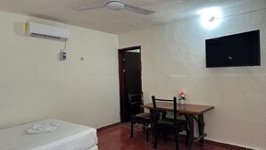 Une petite pièce comprenant un lit, une table avec des chaises, un ventilateur de plafond, un climatiseur et un téléviseur fixé au mur.