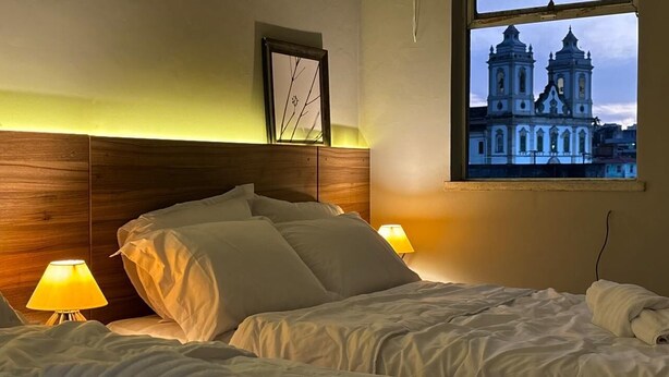 A hotel room with a bed, two bedside lamps, a window with a view of a cathedral, and a framed picture on the wall.
