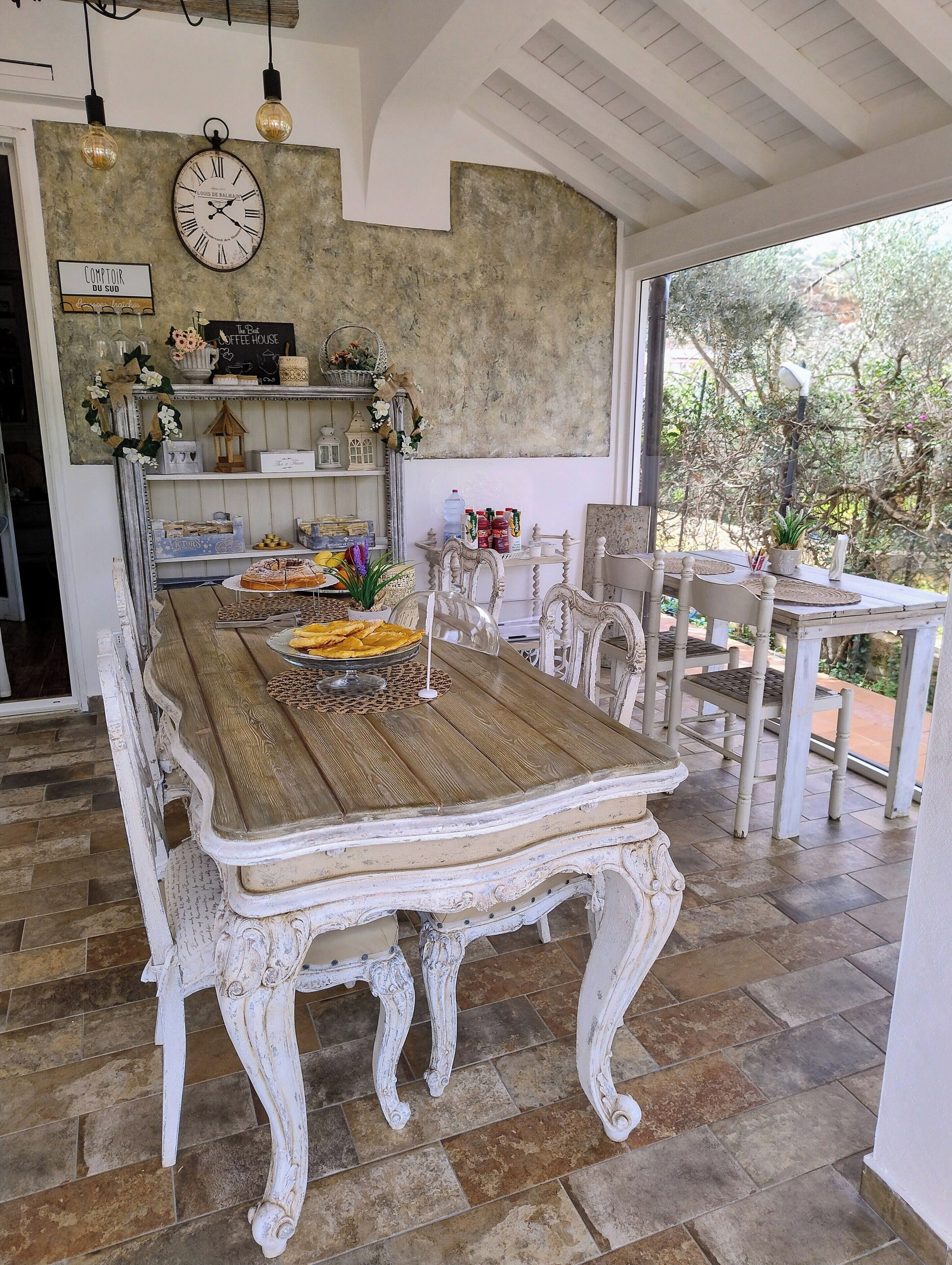 Un comedor rústico con una mesa de madera y sillas blancas decorativas, un reloj en la pared y vistas al exterior.