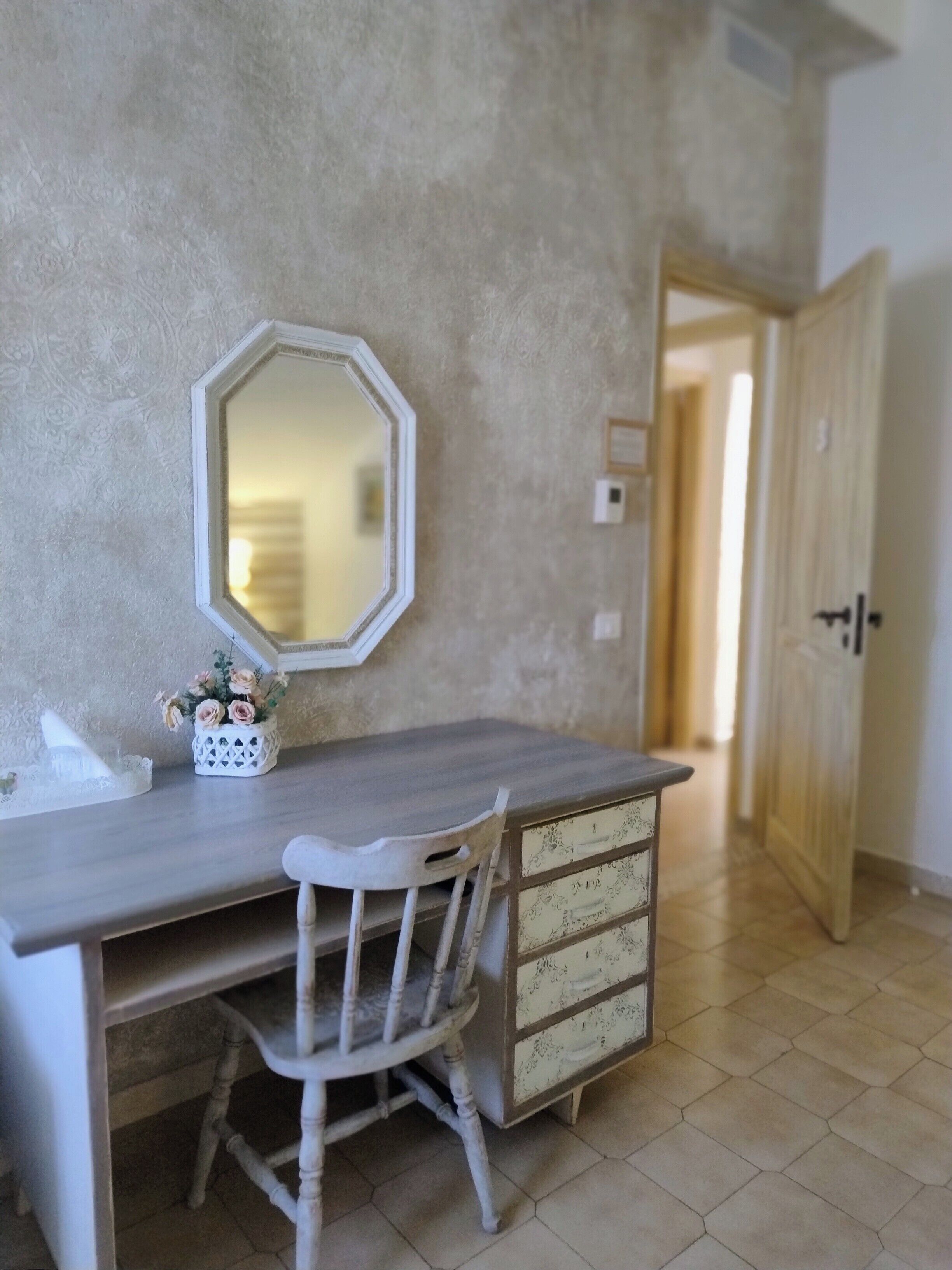 Una habitación con un escritorio de madera, una silla blanca, un espejo decorativo y un aparador con cajones.