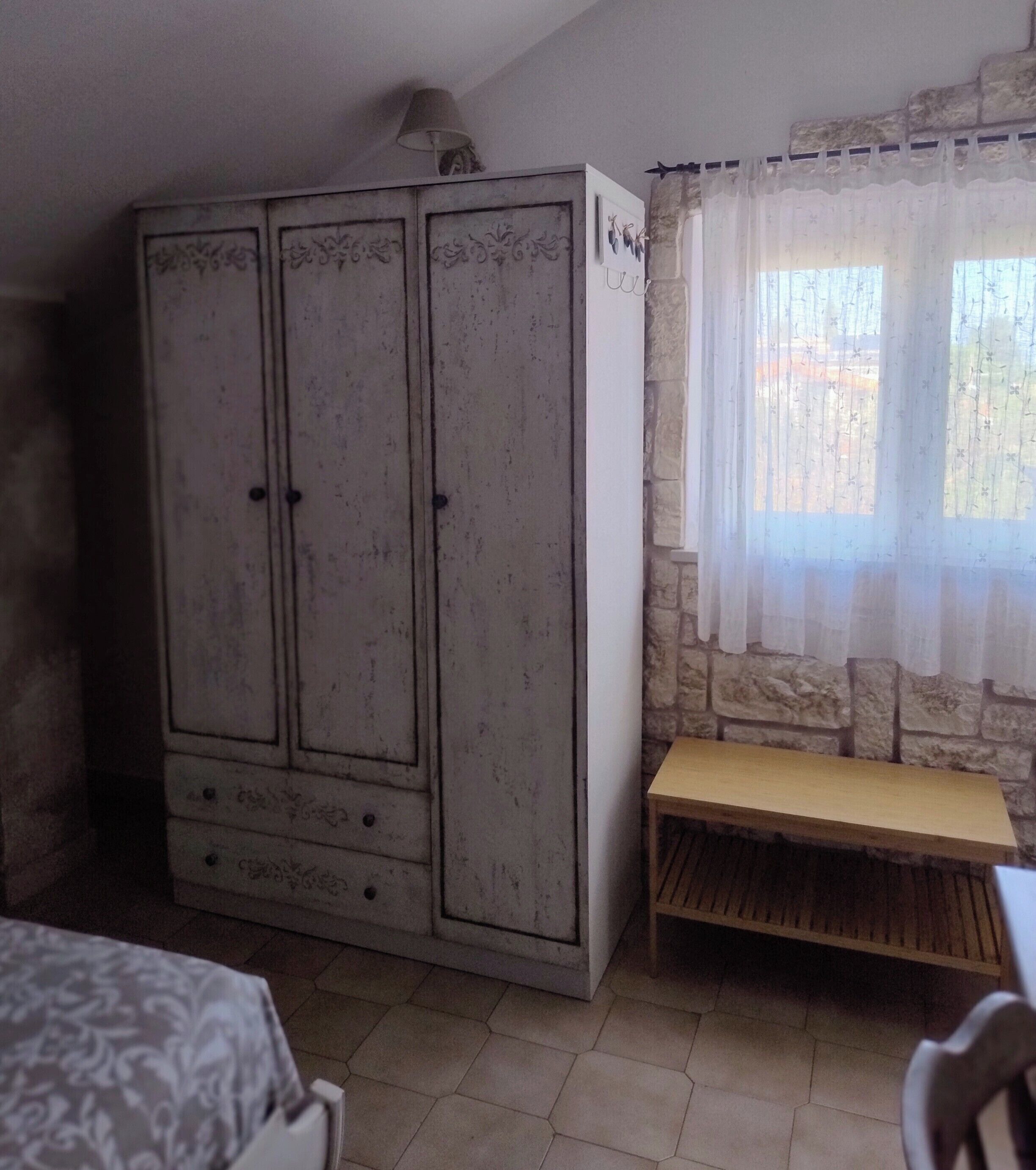 Una habitación con un armario grande, una mesita de madera y una ventana con cortinas traslúcidas.