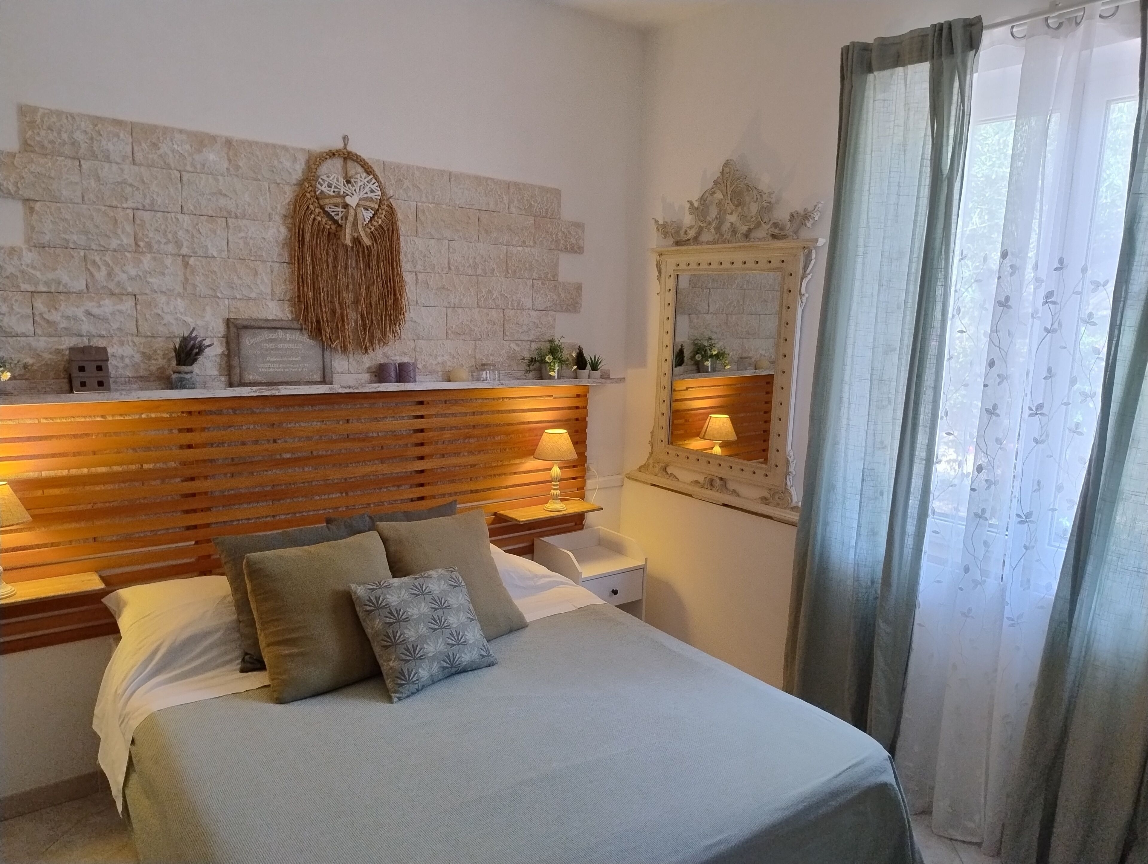 Quarto com parede de pedra, cabeceira de madeira, luminárias de cabeceira, cama com almofadas, mesa de cabeceira, espelho e janela com cortinas.