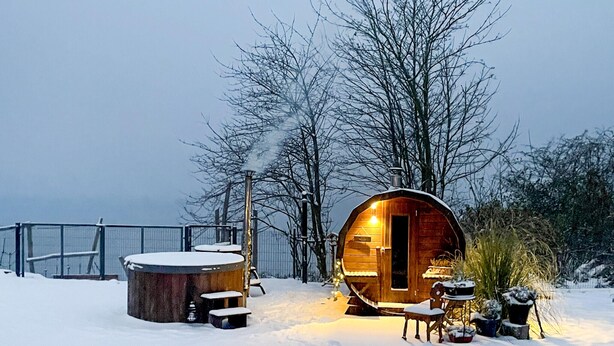 Une cabane en bois, dont l’intérieur est éclairé d’une lumière chaleureuse, entourée d’un paysage enneigé et d’arbres.