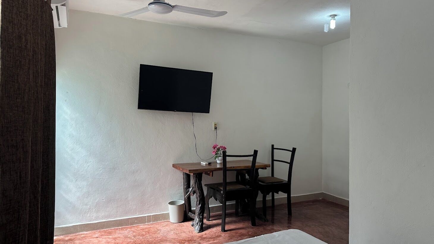 Una habitación con un televisor de pantalla plana montado en la pared, una mesa de comedor de madera con cuatro sillas, un ventilador de techo y un cubo de basura pequeño.