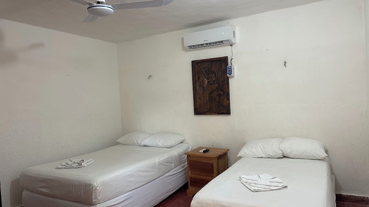 A room with two single beds, a wooden nightstand, a ceiling fan, an air conditioning unit, and a wall-mounted electrical outlet.