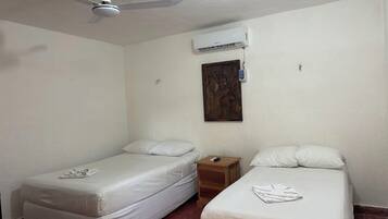 A room with two single beds, a wooden nightstand, a ceiling fan, an air conditioning unit, and a wall-mounted electrical outlet.