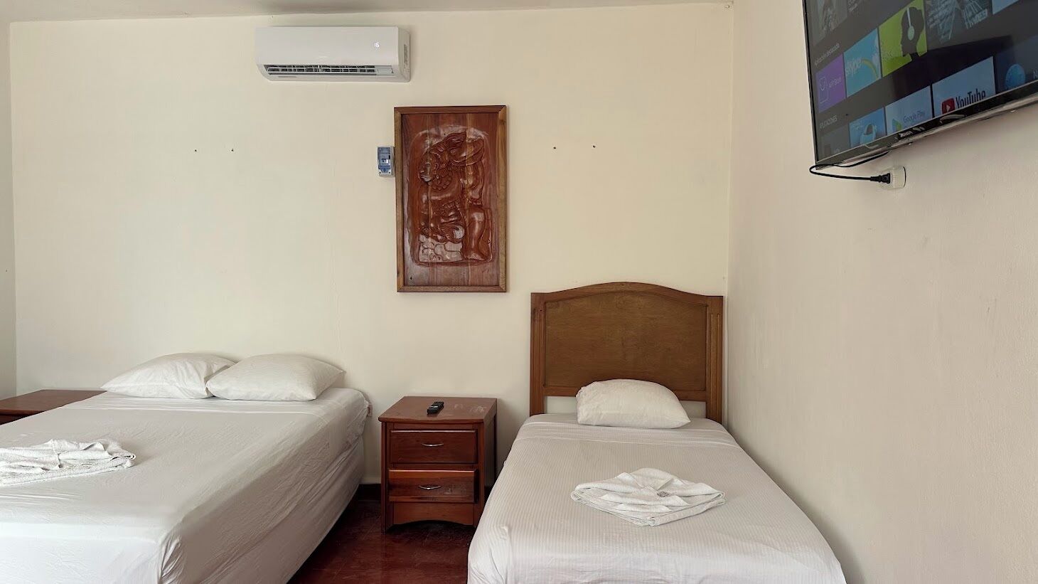 Una habitación de hotel con dos camas individuales, una mesita de noche de madera, un aire acondicionado de pared y una obra de arte enmarcada sobre la cama izquierda.