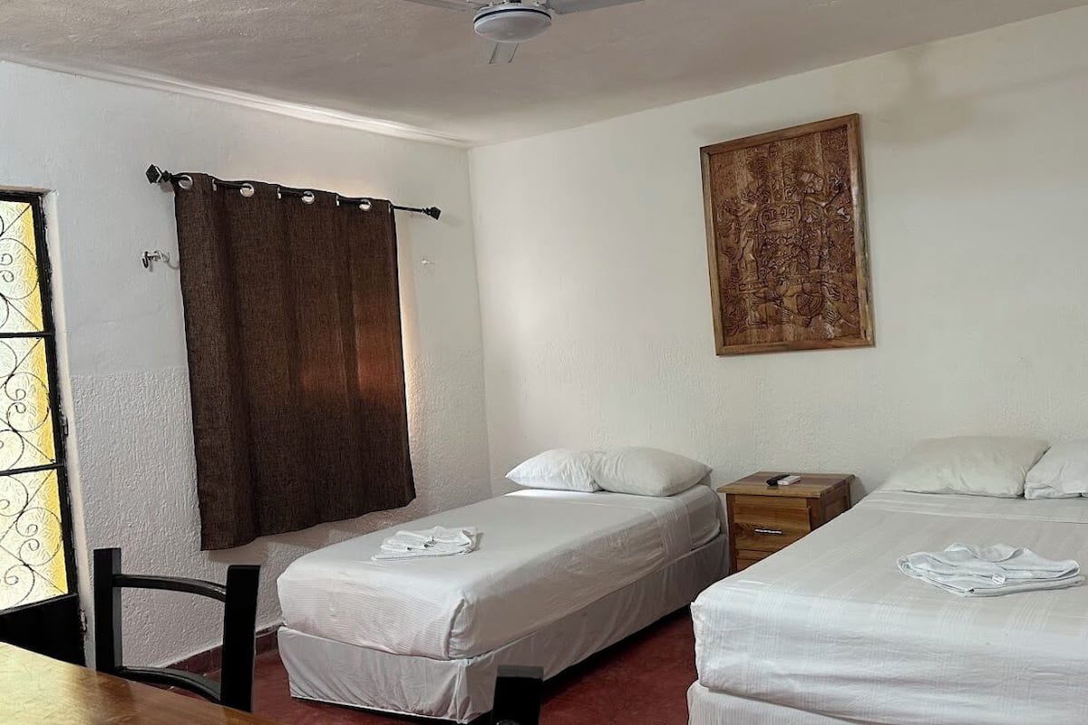 A room with two single beds, a wooden nightstand, a small table, a ceiling fan, and a window with curtains.
