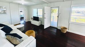 A modern living room with a white sofa, a flat-screen TV, and a large window offering a view of the outdoors. - Playgrounds -Your perfect Family Getaway Awaits! (Lancelin)