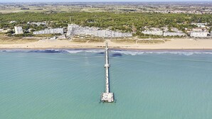 Vista aérea de uma área costeira com um longo cais que se estende para o mar, cercado por edifícios residenciais e uma praia.