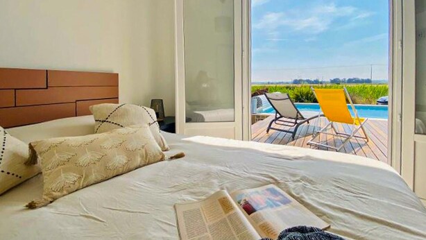 Quarto com uma cama grande, janela com vista para uma piscina e cadeiras ao ar livre, e um livro na cama.
