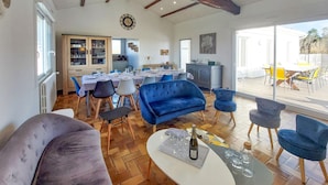 Uma sala de estar ampla, com um sofá azul, uma mesa de centro branca e um espaço de jantar com mesa posta para quatro pessoas.