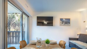 Un coin repas avec une table en bois dressée pour quatre personnes, un téléviseur fixé au mur et un balcon offrant une vue sur les arbres et les bâtiments.