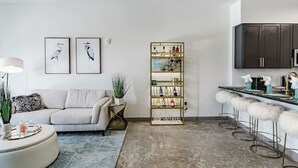 Ein modernes Wohnzimmer mit einer Couch, einem Couchtisch und einer Bar mit Hockern. Ein Bücherregal mit verschiedenen Gegenständen und gerahmten Kunstwerken ist an der Wand zu sehen.