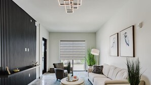 Ein modernes Wohnzimmer mit Sofa, Sessel und Hocker, einer vertikalen Wandlamelle in Schwarz und einem großen Fenster mit Jalousien.