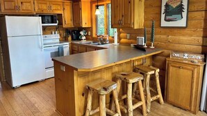A wooden cabin kitchen with a central island, bar stools, a refrigerator, microwave, stove, and a sign reading "S'mores Station".