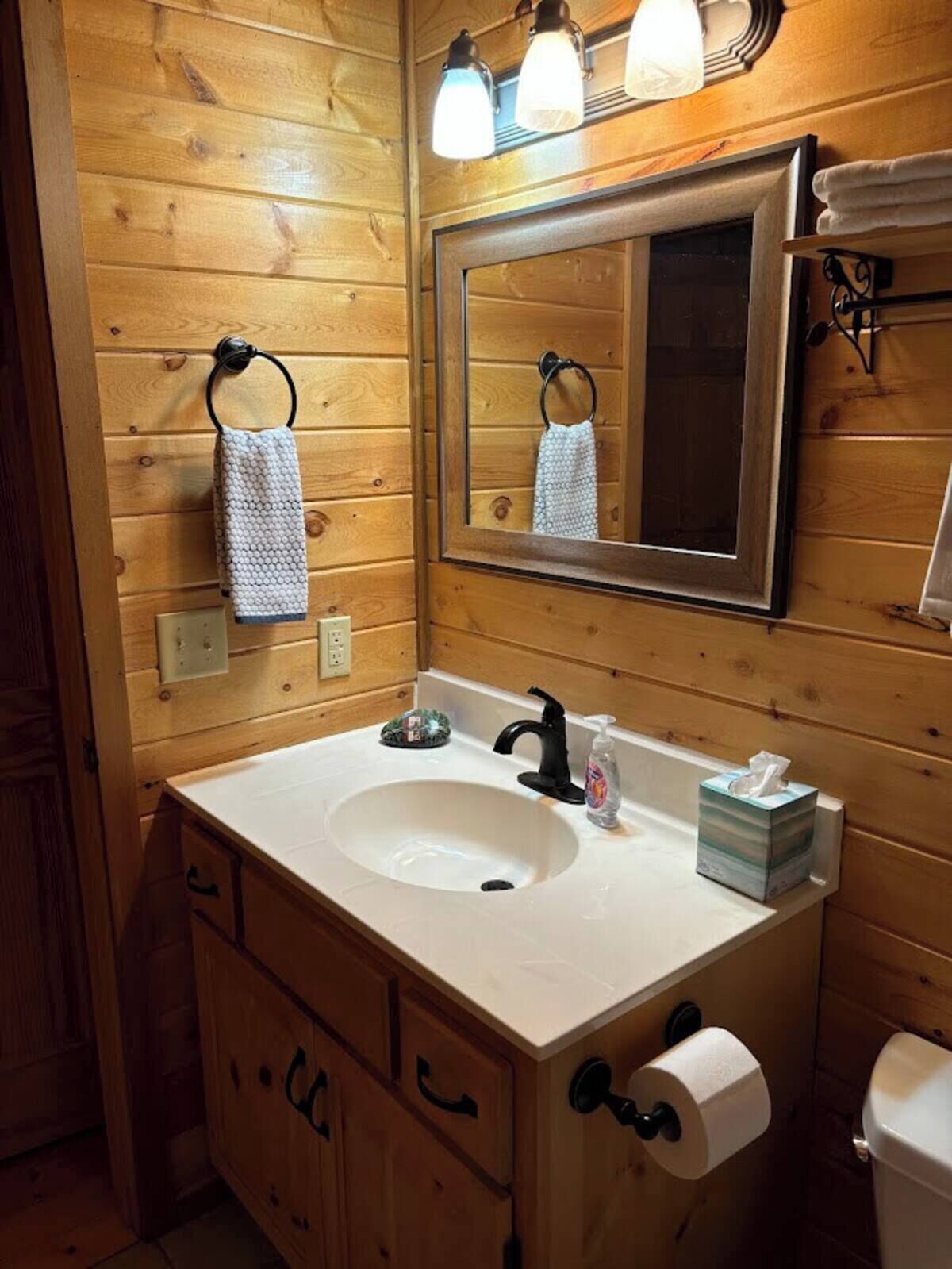 Ein Badezimmer mit Holzvertäfelung, einem weißen Waschbecken, einem schwarzen Wasserhahn und einem Spiegel darüber. Zwei Handtücher hängen an Haken, neben dem Waschbecken befindet sich ein Seifenspender und eine Taschentuchbox.