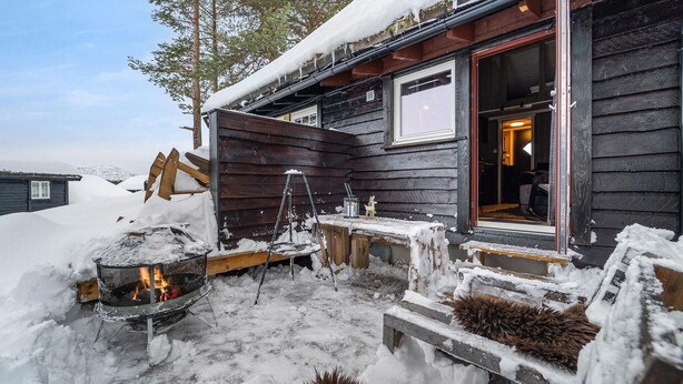 En stuga byggd av trä med en eldstad utomhus, trappsteg som leder upp till ingången och ett litet bord med en stol.