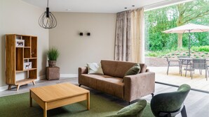 A modern living room with a sofa, coffee table, and chairs. There is a wooden bookshelf with decorative items and a hanging light fixture. A large window offers a view of a garden outside. - 2 bedroom gorgeous home in Afferden (Afferden)