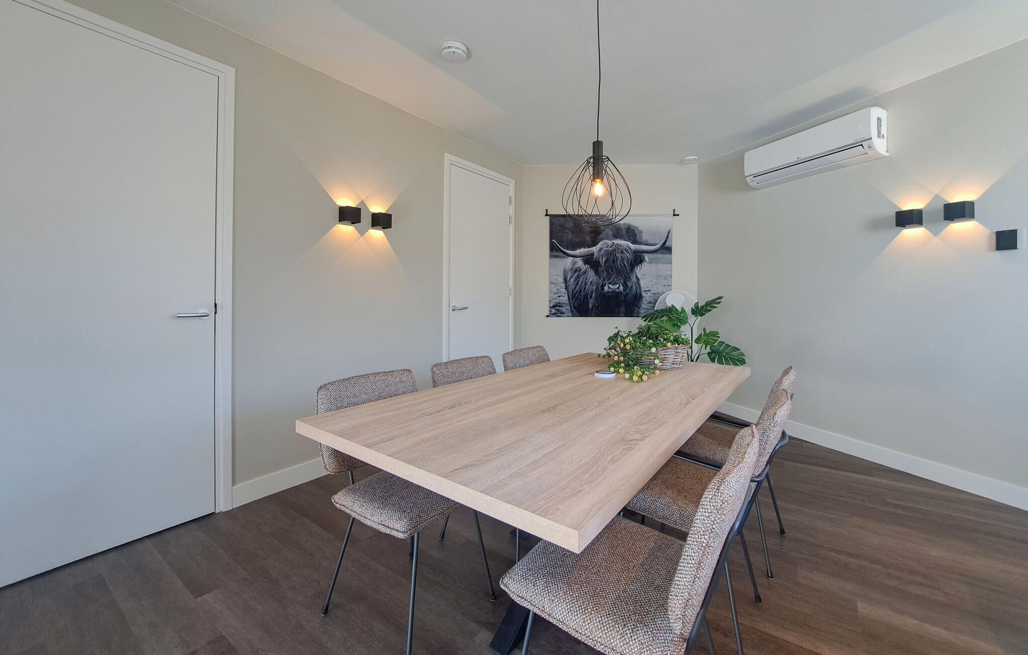 Une salle à manger moderne avec une longue table en bois, six chaises et un luminaire suspendu.