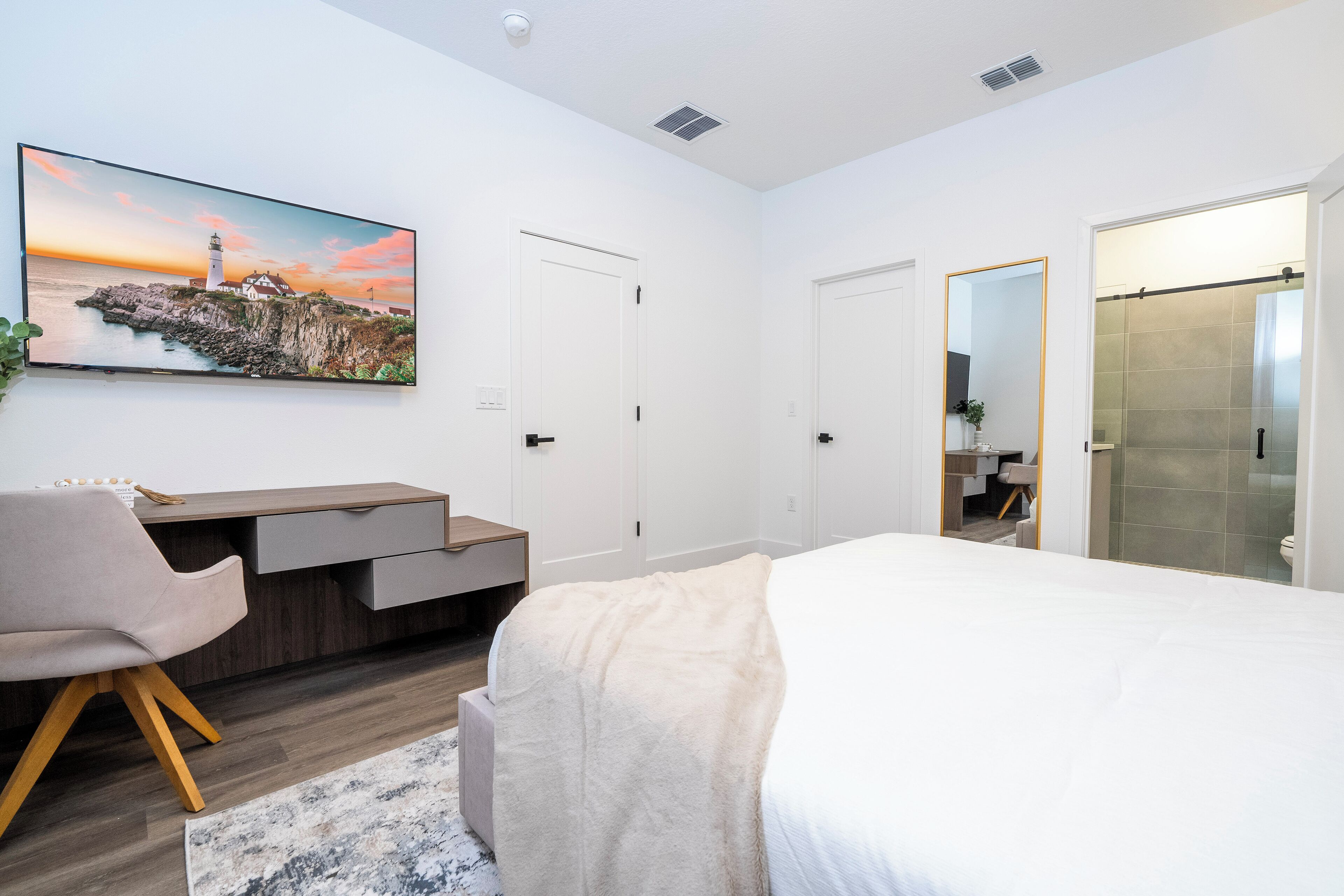 Quarto moderno com uma cama, uma escrivaninha e uma cadeira. Há um quadro na parede e uma porta que dá para o banheiro.