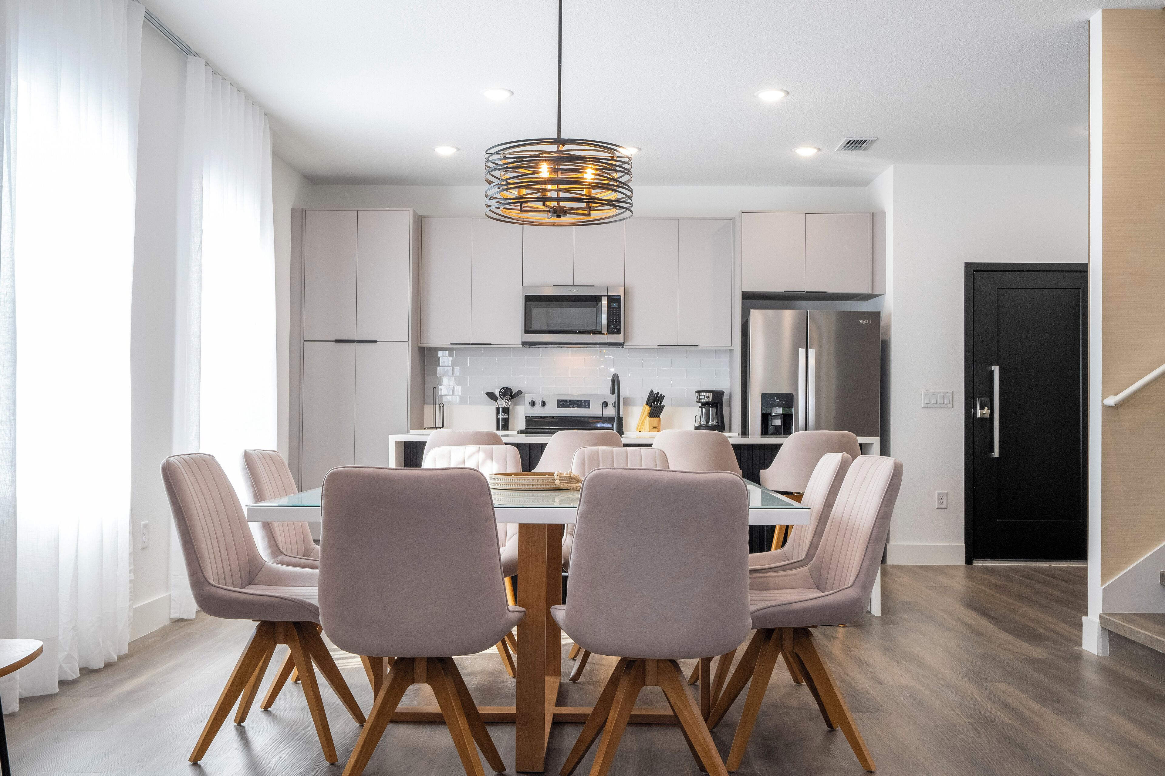 A modern kitchen with stainless steel appliances, a dining area with a glass table and cushioned chairs, and a staircase to the right.
