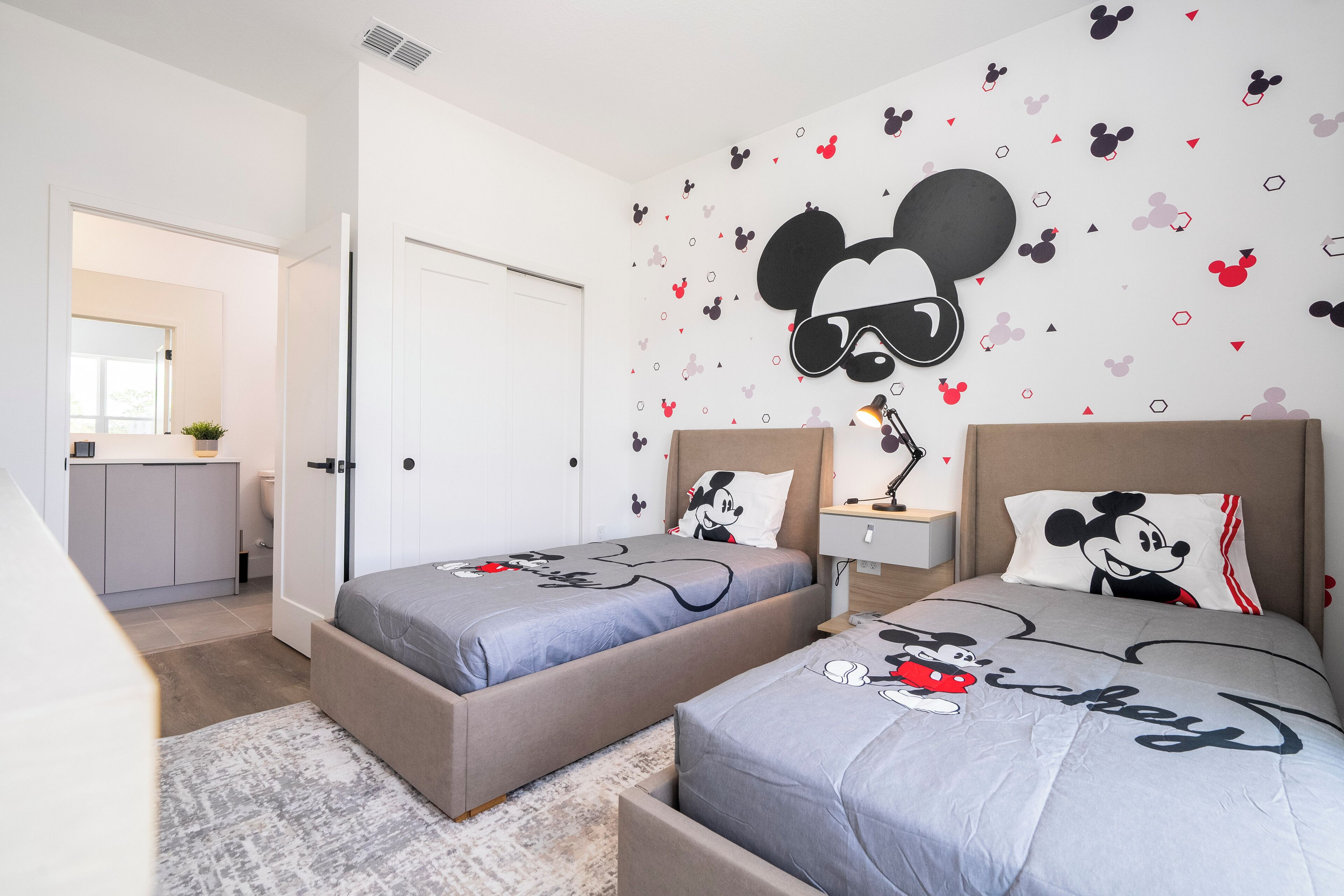 Un dormitorio con dos camas, cada una con ropa de cama temática de Mickey Mouse. Hay un escritorio pequeño con una lámpara y una mesita de noche con una lámpara. La habitación tiene una puerta blanca y una ventana.