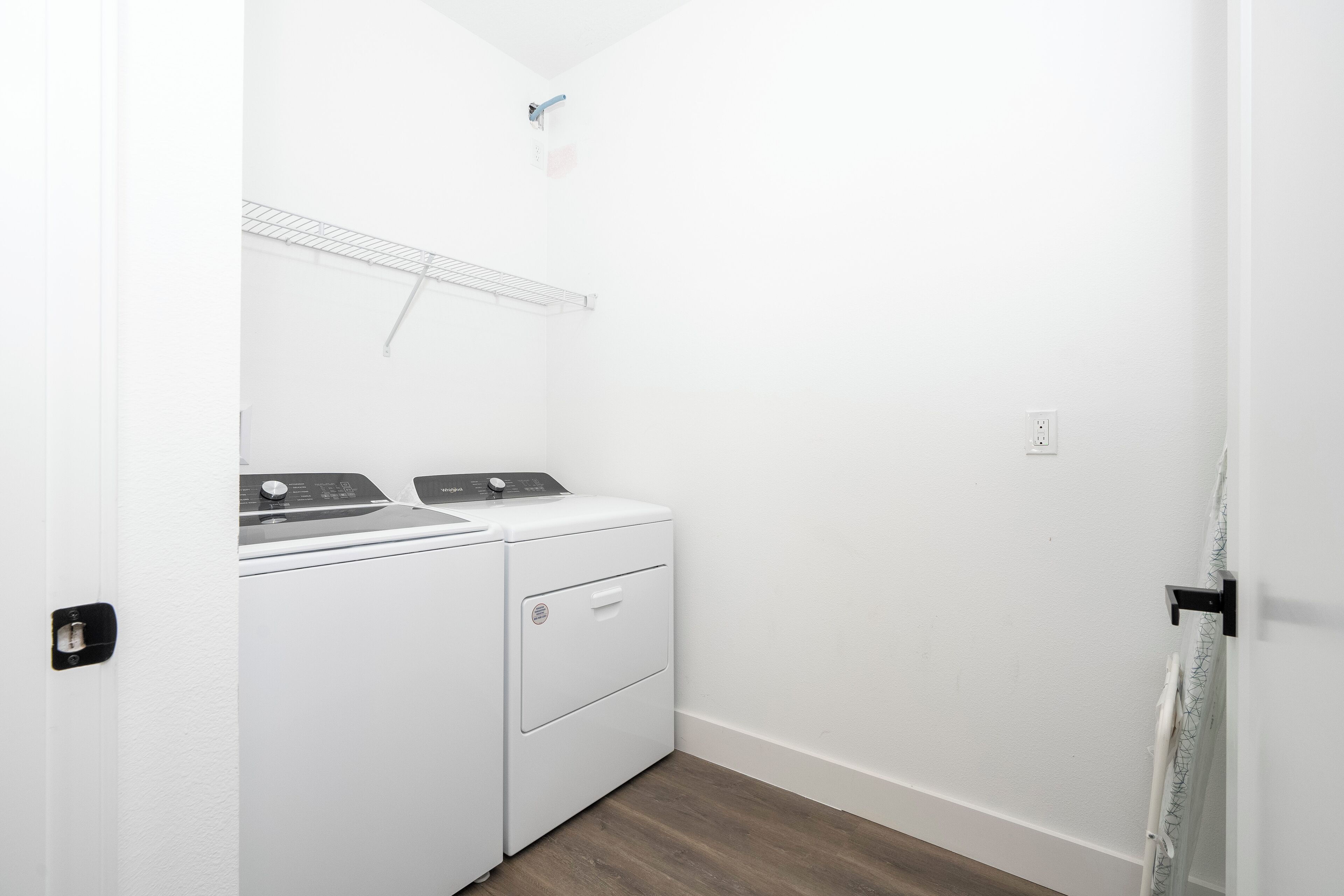 A laundry room with a front-loading washer and dryer, a metal shelf, and a door with a black handle.