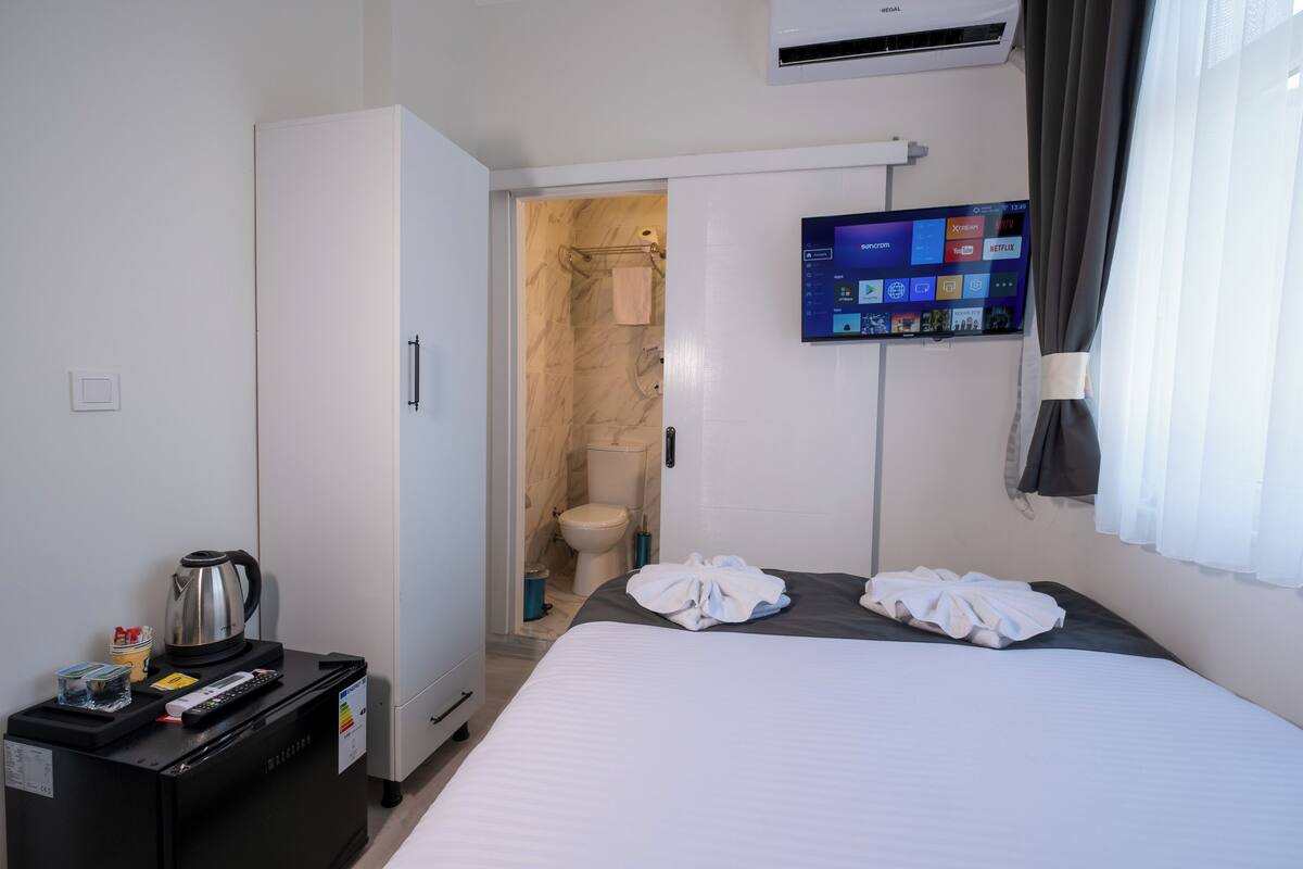 Quarto de hotel com cama, televisão na parede, guarda-roupa, casa de banho com chuveiro e máquina de café num apoio lateral.