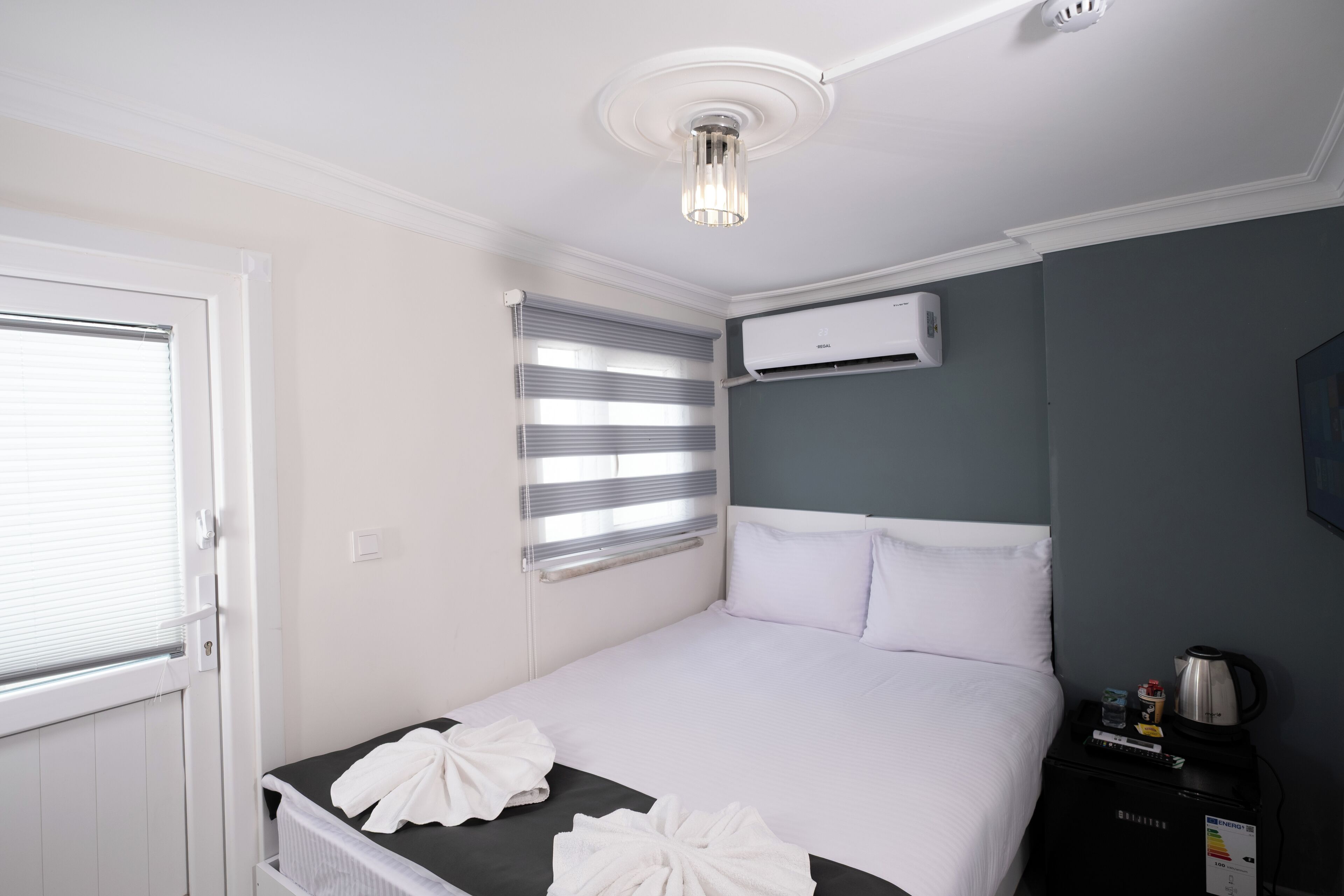 A hotel room with a bed, a radiator, a window with blinds, an air conditioning unit, and a nightstand with a kettle and a bottle.