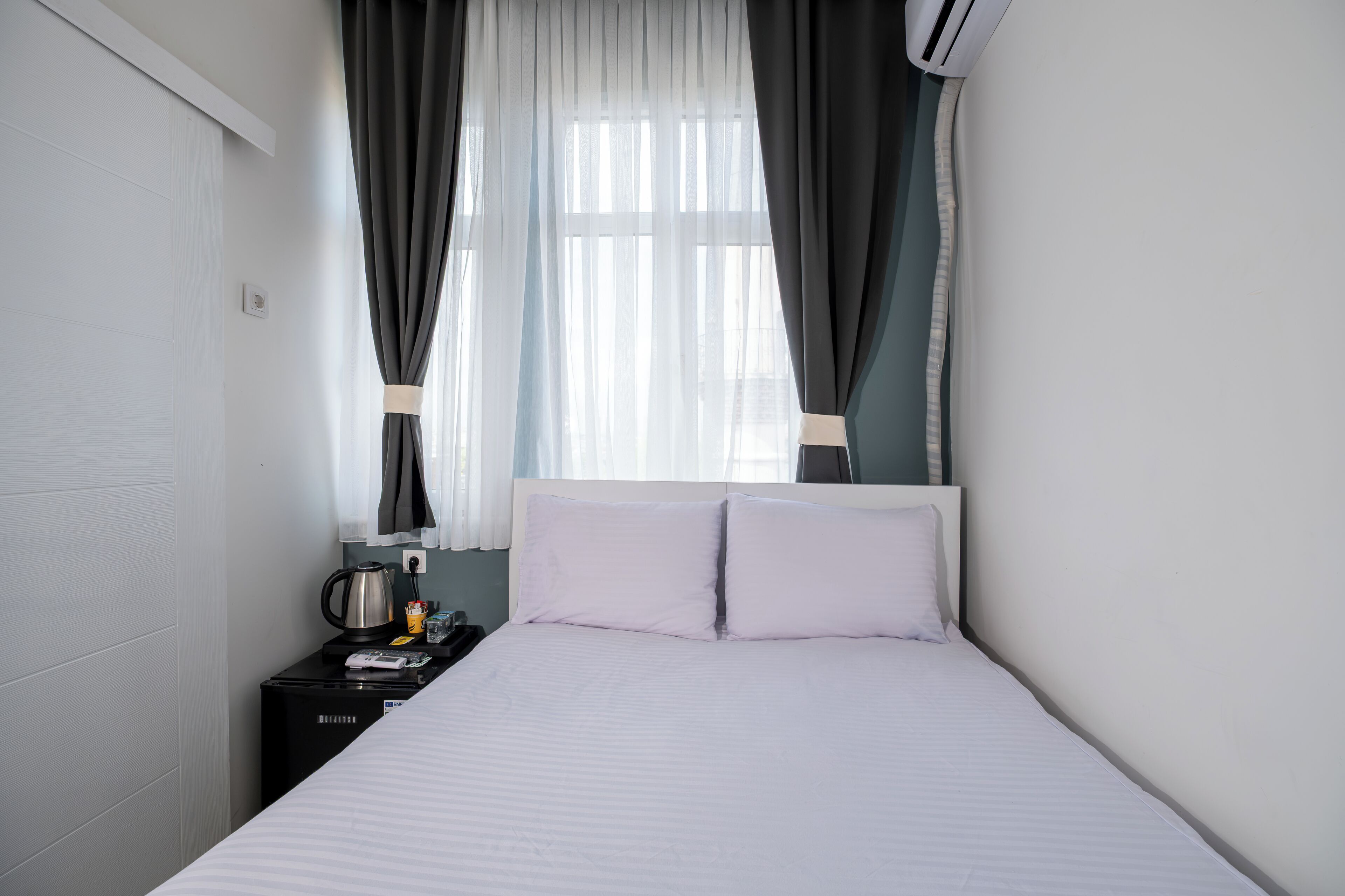 A neatly made bed with white linens, a bedside table with a kettle, and a window with sheer curtains.