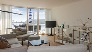 A modern living room with a large window, a flat-screen TV, and a glass shelving unit.