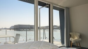 A bedroom with a large window overlooking a body of water and a building. There is a bed with white bedding and a chair with a fluffy cushion.