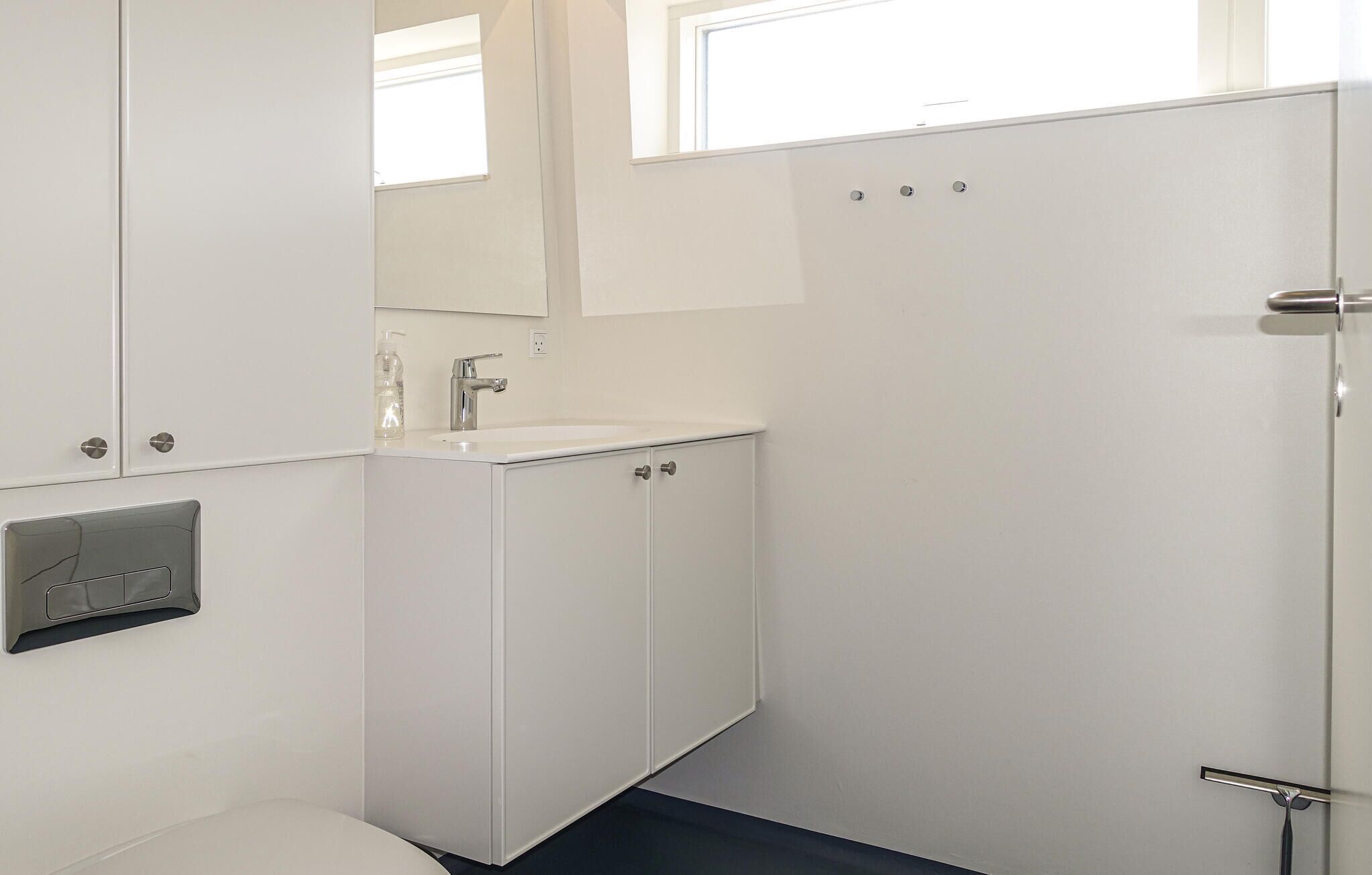 Een badkamer met een wit wastafelmeubel, een spiegel en kastjes. Er is een raam boven de spiegel en een deur aan de rechterkant.
