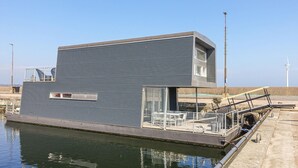 A modern floating house with a flat roof, large windows, and a small deck area with outdoor furniture.