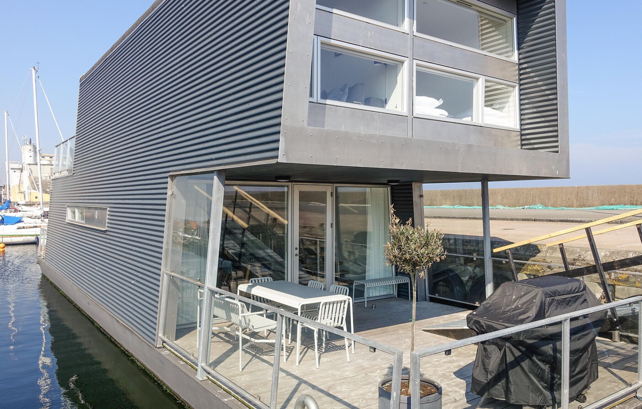 A modern houseboat with a deck, outdoor seating, and a covered grill area.