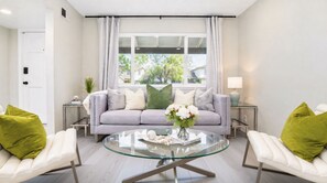 Een moderne woonkamer met een glazen salontafel, een grijze bank met kussens, twee witte fauteuils en uitzicht op een woonwijk door een groot raam.