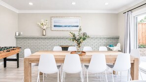 Un comedor con una mesa de madera y sillas blancas, una pared de ladrillo, un cuadro enmarcado y una mesa de futbolín.