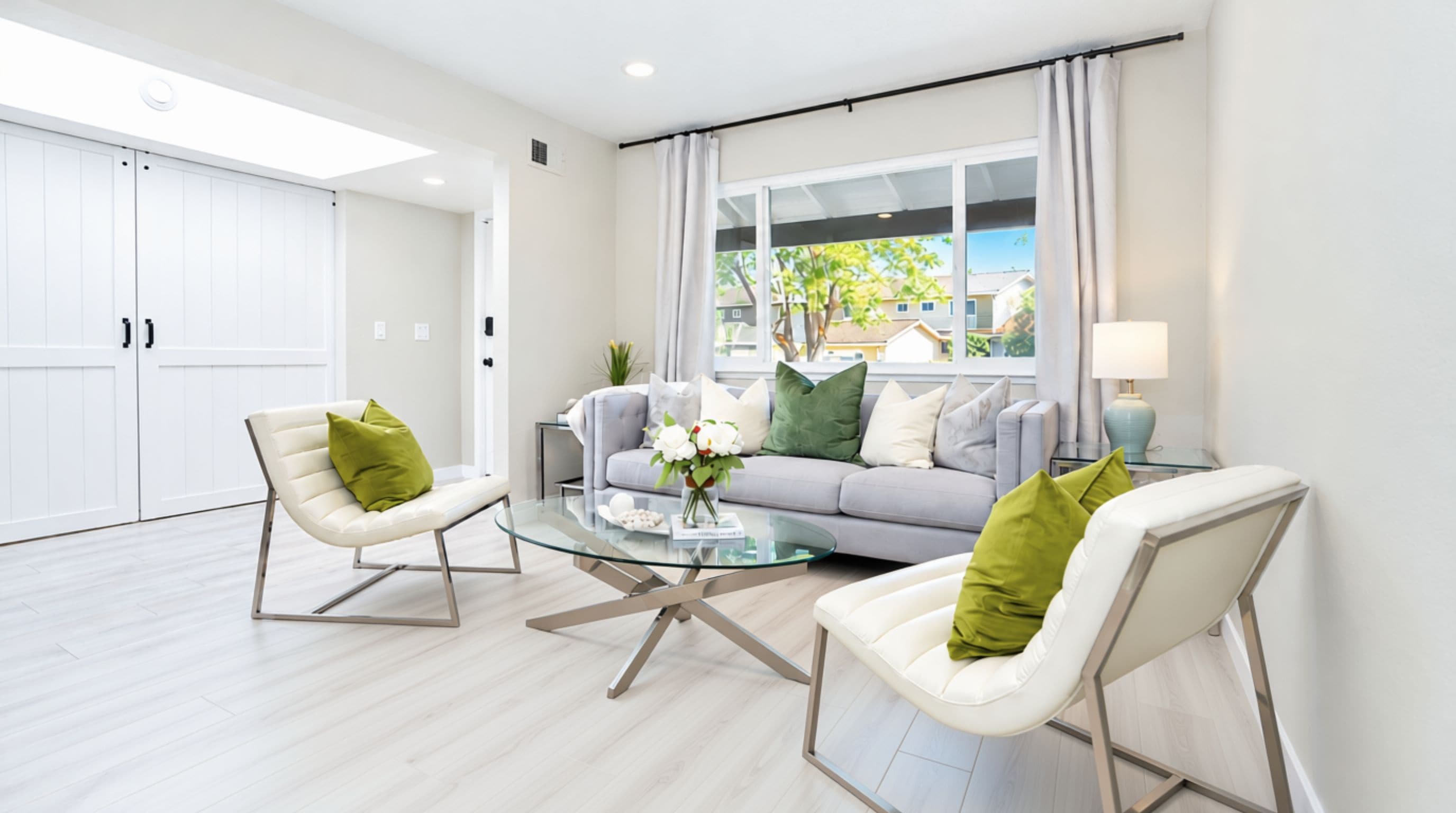 Un salón moderno con una mesa de centro de vidrio, dos sillas blancas con cojines verdes, un sofá gris con varios cojines y vistas al exterior a través de amplios ventanales.