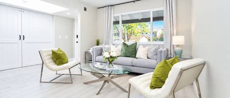 Un salón moderno con una mesa de centro de vidrio, dos sillas blancas con cojines verdes, un sofá gris con varios cojines y vistas al exterior a través de amplios ventanales.