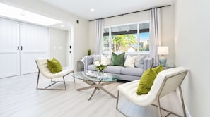 Un salón moderno con una mesa de centro de vidrio, dos sillas blancas con cojines verdes, un sofá gris con varios cojines y vistas al exterior a través de amplios ventanales.