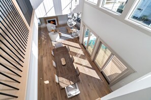 Un vaste espace de vie de style loft, doté d’un plancher en bois, d’un coin repas avec une longue table et des chaises, d’une cheminée et de grandes fenêtres offrant une vue sur l’extérieur.