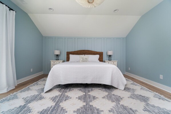 A bedroom with a large bed, two bedside tables with lamps, a patterned rug, and light blue walls. - New luxury home in prestigious Roosevelt Beach (Pine Knoll Shores)