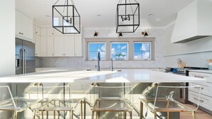 Eine moderne Küche mit einer großen Kochinsel, Barhocker, Pendelleuchten und Blick zum Himmel durch Fenster.