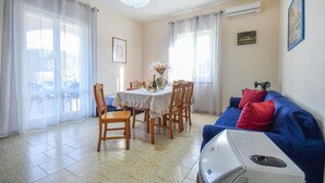 A dining area with a table set for four, surrounded by wooden chairs. There is a blue sofa with red cushions to the right. An air conditioning unit is mounted on the wall, and a framed picture hangs above it.
