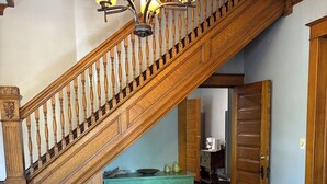 Una escalera de madera con un candelabro, una mesa de comedor y un tapete estampado en una habitación con puertas de madera y pared azul claro.