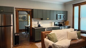 Una cocina moderna con gabinetes grises, un refrigerador de acero inoxidable y un microondas. En la sala de estar, hay un sofá de cuero marrón con una manta y almohadones.