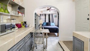 Een compacte keuken met een magnetron, een eettafeltje en een stoel. In de aangrenzende kamer staat een bed met een blauwe dekbedovertrek en een plafondventilator.