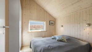 A bedroom with a sloped ceiling, wooden walls, a bed with a grey bedspread, a window with a view of trees, and a small bedside table.
